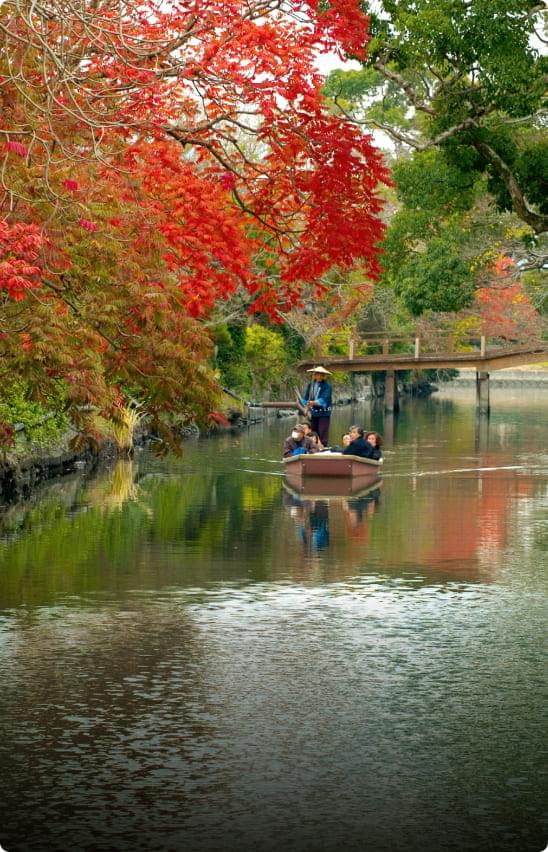 秋の柳川市を彩る風景。紅葉した木々が水辺に映り込み、川沿いの遊歩道には赤や黄色の落ち葉が広がる。どんこ舟が静かに進み、涼しい秋風が水面を揺らす。